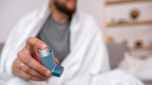 closeup of an asthma inhaler being held by a man in a greay shirt and wrapped in a white blanket